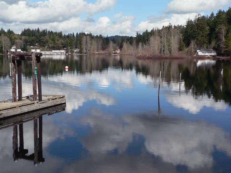 A wooden dock on Shawnigan Lake with the shoreline and clouds reflected in calm water