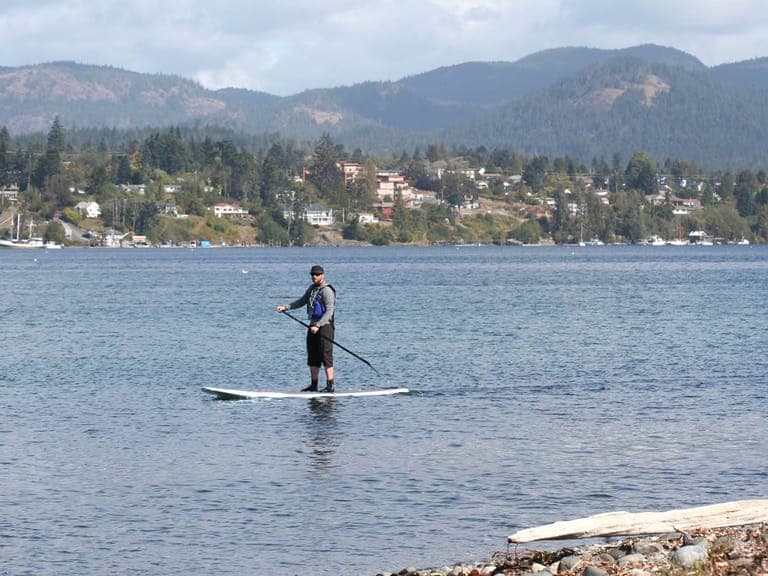 A paddleboarder on the water off Whiffin Spit, with Sooke and the hills behind