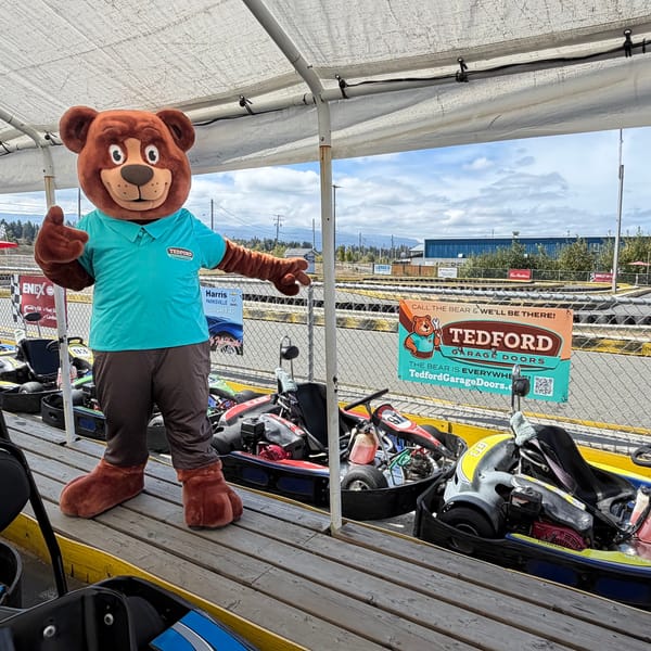 The Tedford bear mascot at a kart track with a Tedford banner on the fence